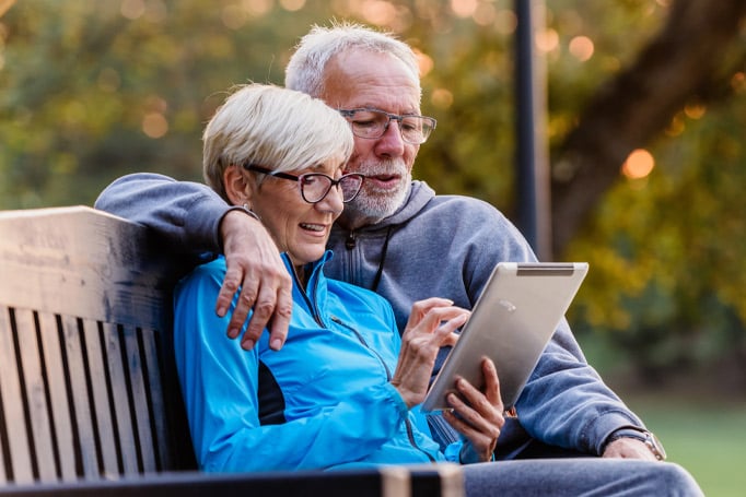 An elder couple wearing jackets sitting on a park bench looking at their tablet